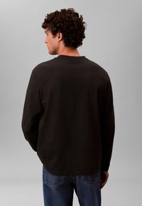 Hombre con cabello rizado de espaldas, vistiendo una camisa negra de manga larga y jeans azules sobre un fondo gris liso.