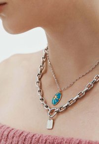 Collar de cadena en tono plateado, superpuesto con un colgante de turquesa que presenta un diseño en forma de corazón y una pequeña etiqueta rectangular. Se pueden ver los eslabones texturizados.