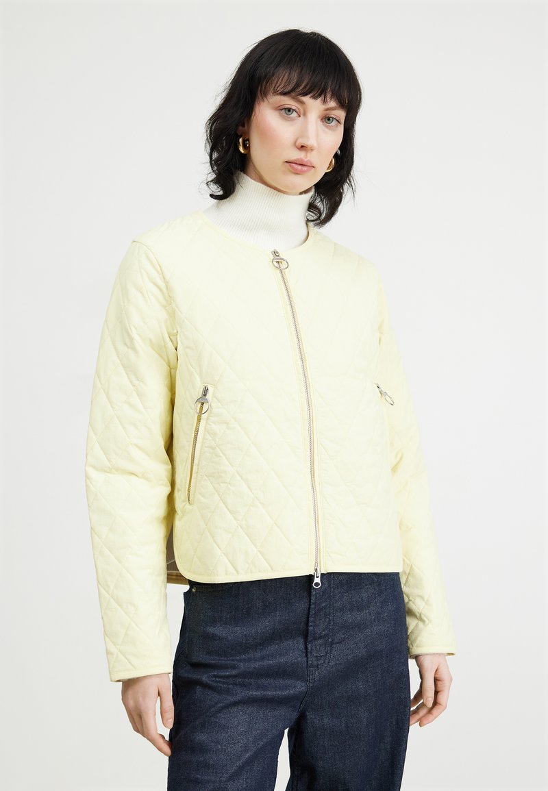 Gesteppte gelbe Jacke mit einem kurzen Design, zwei mit Reißverschlüssen versehenen Taschen und einem silbernen Reißverschluss auf der Vorderseite. Getragen über einem weißen Rollkragenpullover.