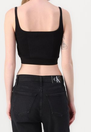 Kvinde iført en sort ribbet crop top og sorte højtaljede jeans med et Calvin Klein-logopatch på linningen, set bagfra.