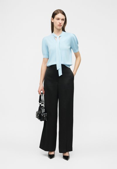 Η γυναίκα φοράει γαλάζιο μπλουζάκι με κοντά μανίκια και γραβάτα στο λαιμό, μαύρο φαρδύ παντελόνι, μαύρα μυτερά παπούτσια και κρατά μια μικρή μαύρη τσάντα.