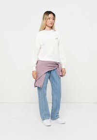 Hvid skjorte med lange ærmer og et lille logo, løse blå stribede jeans og et lyse lilla tekstureret bånd bundet om taljen; hvide sneakers.