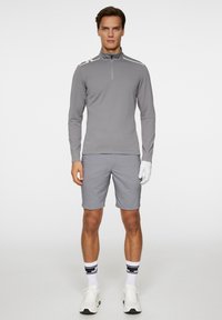 Grå långärmad zip-up skjorta med vita accenter, matchad med grå shorts. Modellen har på sig vita sneakers och en golfhandske, tillsammans med randiga strumpor.