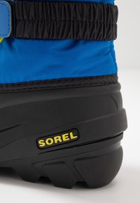 Modrá voděodolná bota s černou gumovou podrážkou; má texturovaný povrch a na boku je žluté logo "SOREL". Viditelné detaily s elastickým páskem.