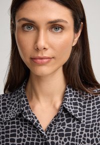 Vrouw met lang bruin haar, blauwe ogen, in een grijs en zwart gestreept overhemd met kraag, kijkt recht vooruit met een neutrale uitstraling.