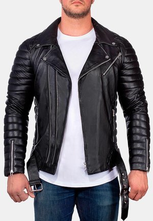 Mann trägt schwarze gesteppte Leder-Bikerjacke mit mehreren Reißverschlüssen über weißem T-Shirt und blauen Jeans, Hände entspannt an den Seiten.