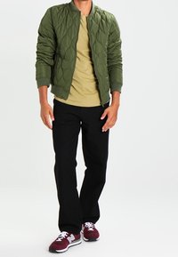 Gequilted olijfgroen bomberjack, mosterdkleurig shirt, zwarte broek en bordeauxrode sneakers met witte veters. Casual, fitted stijl.