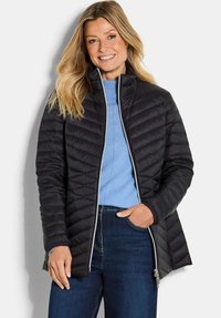 Smiling woman with long blonde hair wearing a black quilted jacket, light blue sweater, and dark blue jeans, standing against a white background.