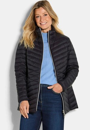 Glunderende vrouw met lang blond haar, gekleed in een zwarte gewatteerde jas, een lichtblauwe trui en donkerblauwe spijkerbroek, staande tegen een witte achtergrond.