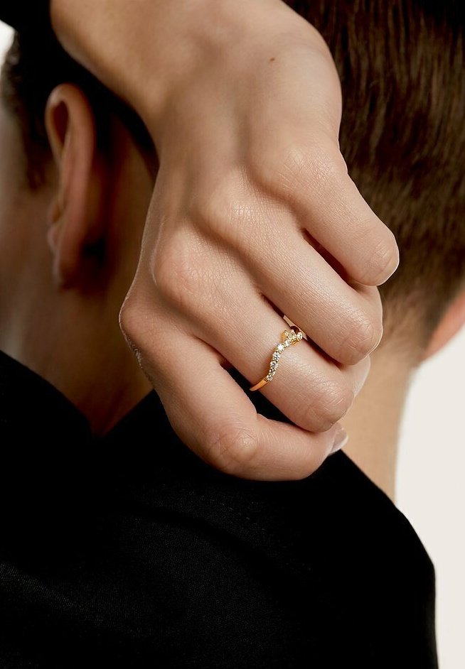 Hånd med en guldring med små diamanter, der hviler på bagsiden af en persons nakke med kort brunt hår og sort tøj.