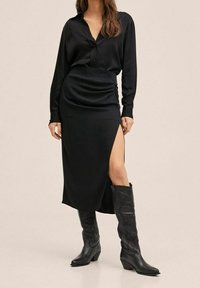 Blusa de satén negra con cierre de botones, combinada con una falda midi negra que presenta una abertura lateral. Llevada con altas botas de vaquero negras.