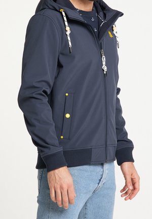 Homme portant un sweat à capuche zippé bleu marine avec des cordons blancs et des boutons jaunes, associé à un jean bleu clair.
