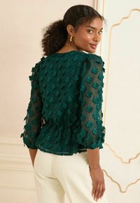 Top verde trasparente con maniche a tre quarti, caratterizzato da un motivo floreale testurizzato e un punto vita peplum. La vista posteriore mostra dettagli a volant sulle spalle.