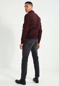 Jachetă bomber burgundy cu manșete și tiv din rib, buzunare cu fermoar, asociată cu blugi negri și bocanci negri la gleznă, pe un fundal alb.