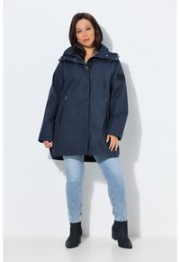 Donna in piedi contro un muro bianco, indossa un cappotto con cappuccio blu navy, jeans azzurri e stivaletti neri, rivolta in avanti con le mani rilassate.