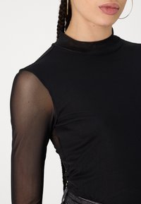 Svart langermet topp med høy hals, glatt solid kropp og gjennomsiktige mesh-ermer med teksturfinish. Minimalistisk design.