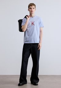 Ανοιχτό μπλε βαμβακερό μπλουζάκι με κόκκινο λογότυπο, συνδυασμένο με σκούρο τζιν παντελόνι με φαρδιά μπατζάκια και μαύρα παπούτσια, κρατώντας μια μαύρη τσάντα tote.