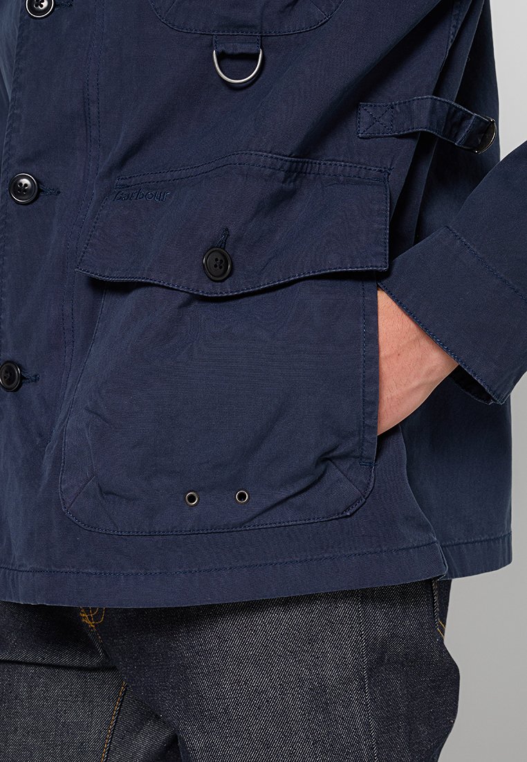 Giubbotto leggero blu navy con tasca con bottone, dettaglio in anello metallico e cinturino sulle maniche. Tessuto morbido con cuciture a contrasto.