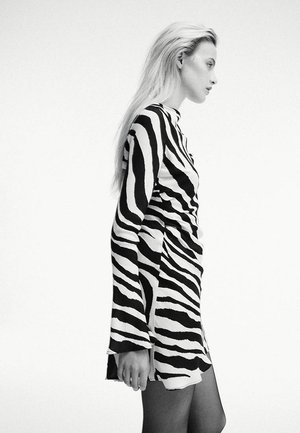 Femme aux longs cheveux blonds portant une robe à rayures zèbre noire et blanche, debout de profil sur un fond clair uni.