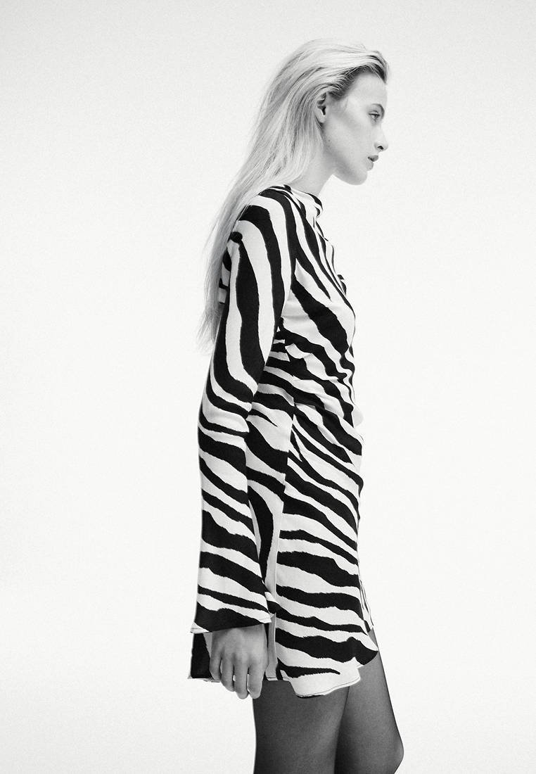Femme aux longs cheveux blonds portant une robe à rayures zèbre noire et blanche, debout de profil sur un fond clair uni.