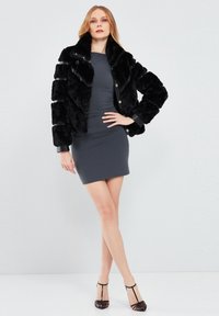 Black faux fur jacket with a collar, featuring a boxy design and button closure. Worn over a fitted gray dress, creating a layered look.