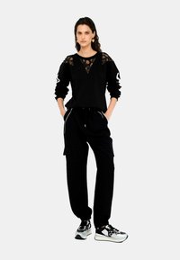 Felpa nera con dettagli in pizzo sulle spalle, abbinata a pantaloni jogger neri con tasche laterali e piping bianco. Sneakers nere e argento.