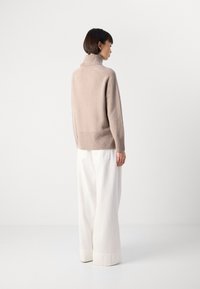 Donna in piedi di profilo, che indossa un maglione beige a collo alto e pantaloni bianchi a gamba larga, su uno sfondo bianco semplice.