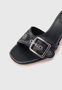 Fekete papucs texturált pánttal, szegecsekkel díszítve, ezüst négyzet alakú csattal, sima bőr talpbéléssel és fehér varrással.