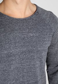 Person som bär en grå stickad tröja med rund halsringning, som visar nyckelbenet och övre bröstområdet.