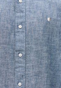 Μπροστινό μέρος μπλε πουκαμίσου chambray με λευκά κουμπιά και μικρό ραμμένο έμβλημα ασπίδας στη δεξιά πλευρά.