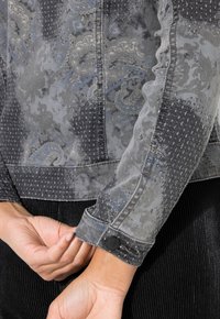 Grå denimjacka med intrikat paisleymönster, texturerad sömnad och svarta knappdetaljer. Närbild som framhäver ärm- och fålldesign.