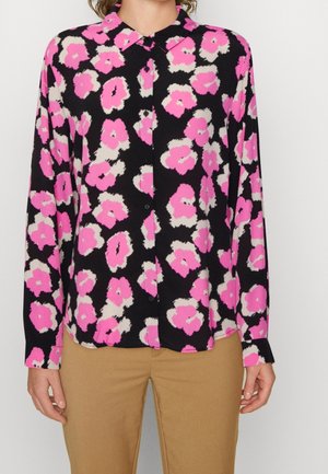 Koszula na guziki z długim rękawem o czarnym tle z jaskrawym różowo-białym abstrakcyjnym kwiatowym wzorem, noszona z beżowymi spodniami.