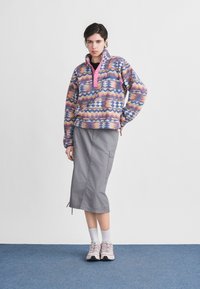 Γυναίκα που φοράει πολύχρωμο φλις πουλόβερ με σχέδια, γκρι μίντι φούστα με πλαϊνή τσέπη, λευκές κάλτσες και χοντρά αθλητικά παπούτσια, στέκεται σε μπλε χαλί.