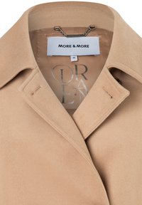 Beige jas van een zachte, gestructureerde stof met een gelapt kraag. Bevat een merklabel en een kettingdetail binnen de halslijn. Maat 36.