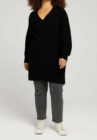 Frau in einem langen schwarzen V-Ausschnitt-Pullover, grauen Jeans und weißen High-Top-Sneakern, die vor einem einfarbigen Hintergrund steht.