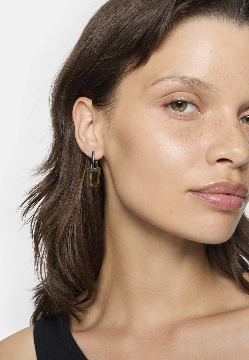 Boucles d'oreilles rectangulaires en or avec une finition brillante et un accent noir mince. Un modèle présente un gros plan, mettant en valeur les détails des boucles d'oreilles.