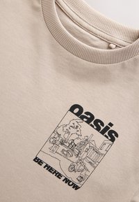 T-shirt beige en coton avec encolure côtelée, arborant une impression graphique noire représentant une scène avec des personnages et des objets, étiquetée "oasis" et "SOIS ICI MAINTENANT".