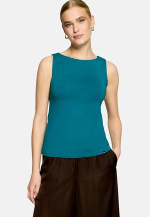 Femme aux cheveux blonds portant des boucles d'oreilles anneaux dorés, un haut sans manches bleu sarcelle et un pantalon noir large, posant avec une main dans la poche.