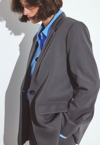 Blazer noir ajusté avec un bouton unique, associé à une chemise bleu clair. Tissu lisse et design structuré, avec poches latérales.