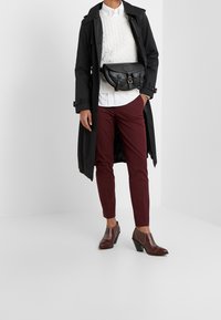 Černý trenčkot, bílý svetr s pleteným vzorem, kaštanově červené tapered kalhoty, černá kožená crossbody taška, hnědé kotníkové boty na podpatku s žebrovanou texturou.