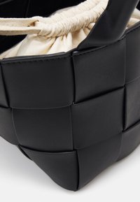 Svart vävd handväska med en texturerad läderutsida och en beigefärgad dragsko av tyg som foder. Har stabila handtag och en strukturerad form.