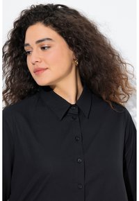 Black button-up shirt featuring a structured collar, smooth fabric, and classic button closure. Curly hair and neutral makeup visible.