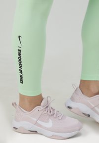 Jasnozielone legginsy mają grafikę "I SWOOSH BY NIKE" w czerni. Różowe buty sportowe z białymi akcentami i teksturowanym wzorem.