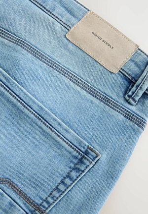 Lys blå denim jeans har en tekstureret overflade, kontrasterende syninger og et gråt stofmærke med "DENIM SUPPLY" præget.
