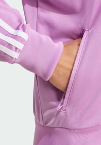 Hand delvis innanför den dragkedjade fickan på en rosa träningsjacka med vita ränder på manschetten.