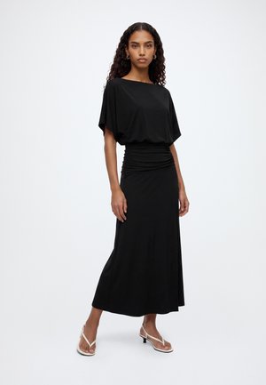 Vrouw draagt een zwarte midi-jurk met een geplooide taille, losse korte mouwen en witte sandalen met hakken, staand tegen een effen achtergrond.