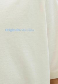 Gaiši bēšas īsās piedurknes T-krekls no mīksta kokvilnas, ar zilu tekstu "Originals Studio Edition" kreisajā krūšu daļā.