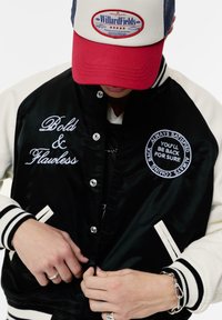 Veste universitaire noire et crème avec texte brodé et écussons, boutons-pression, poignets rayés, et une personne fermant la fermeture.