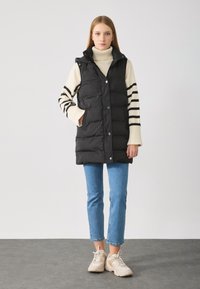 Schwarze Steppweste mit Druckknöpfen, ausgestattet mit einem hohen Kragen und Seitentaschen, getragen über einem gestreiften Pullover und kombiniert mit blauen Jeans und Sneakers.