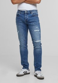 Slim-fit siniset farkut, joissa on kuluneita kohtia, joskus näkyviä maalisuihkeita, viisi taskua ja napitettava sulku, yhdistettynä valkoisiin lenkkareihin.